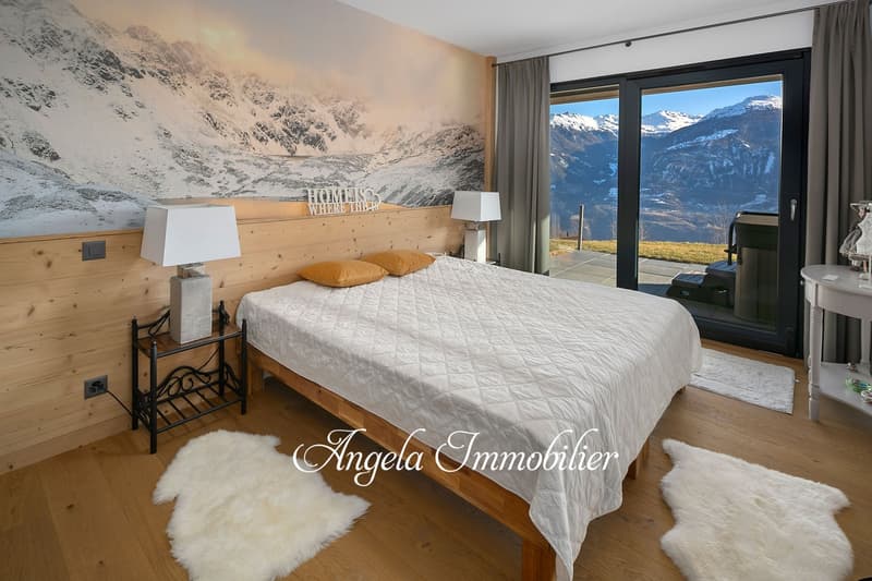 Spacious bedroom with wooden walls and floor, large windows offering a scenic mountain view, a king-size bed with a quilted comforter, and a decorative sign reading 'Home is where the heart is'