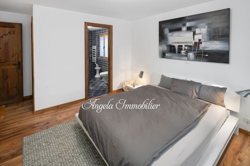 Bedroom with bed, nightstand, lamp, rug, wooden floor, white walls, wooden door, abstract painting, bathroom with toilet and window