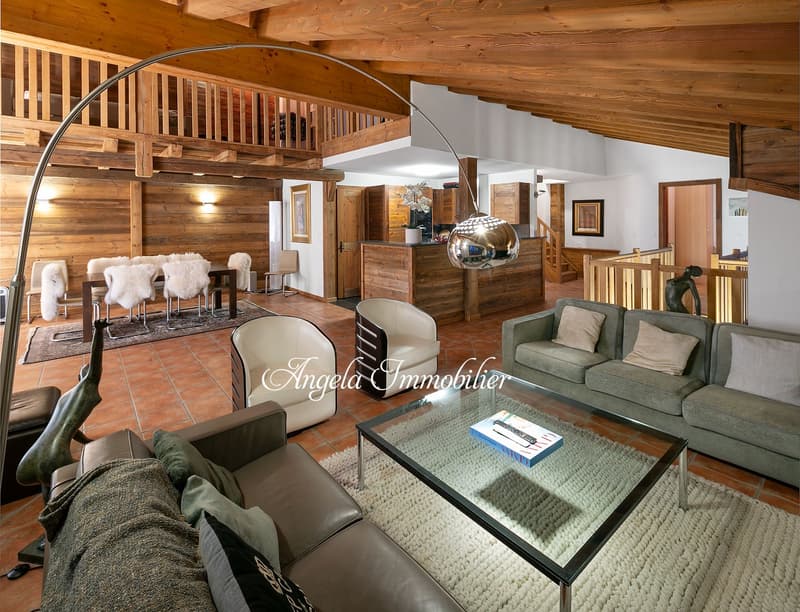 Spacious open-plan living area with wooden beams and walls, high ceilings, a cozy fireplace, comfortable sofas and armchairs, a glass coffee table, and a dining area with chairs and a table.