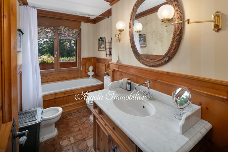 Spacious bathroom with wooden paneling, large round mirror, vanity with marble countertop, and a bathtub with a view of the outdoor garden through the window.