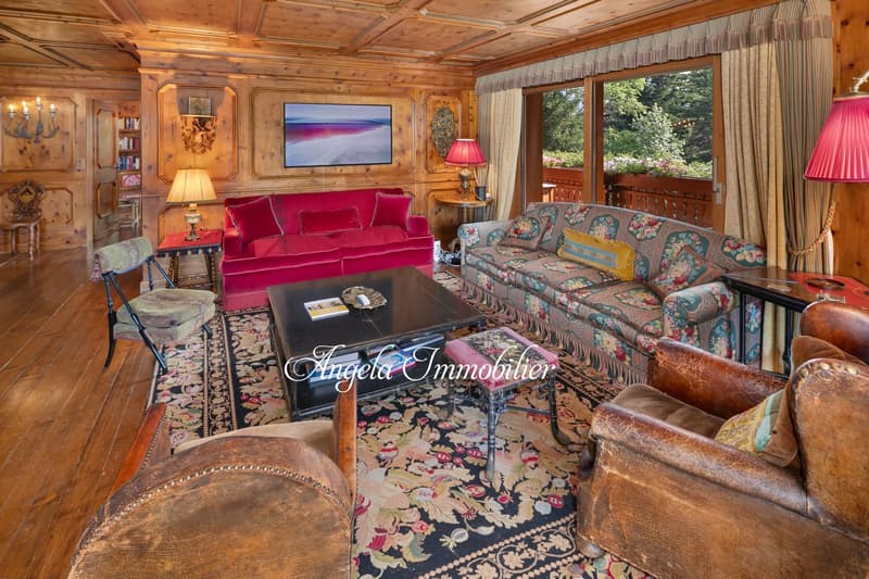 Cozy and rustic living room with wooden walls and ceiling, large red velvet sofa, floral patterned armchairs, wooden coffee table, fireplace, and large windows overlooking the outdoor scenery