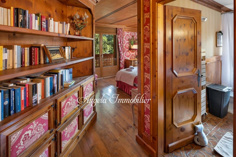 Cozy bedroom with wooden walls and shelves, pink patterned curtains, a bed, and a fireplace. The room also has a bookshelf filled with books.