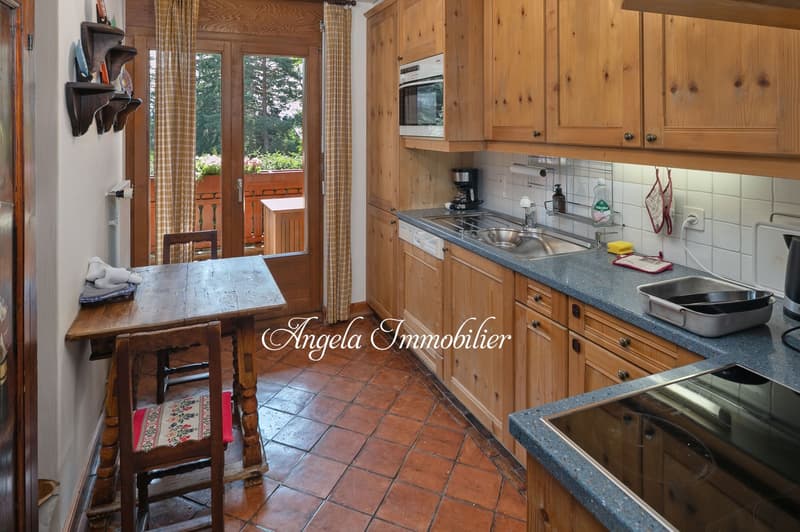Rustic kitchen with wooden cabinets, tile flooring, large windows overlooking a balcony or terrace, and a dining table with chairs