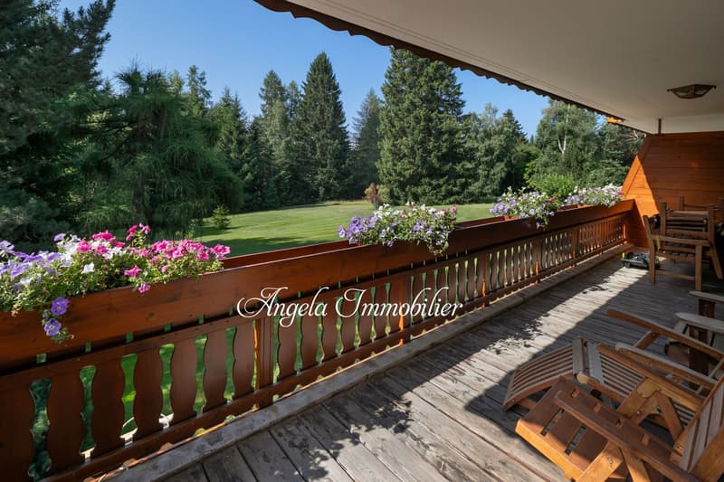 Wooden deck with railing, surrounded by lush greenery and pine trees, overlooking a grassy lawn. Flowers in planters along the railing. Wooden furniture on the deck.