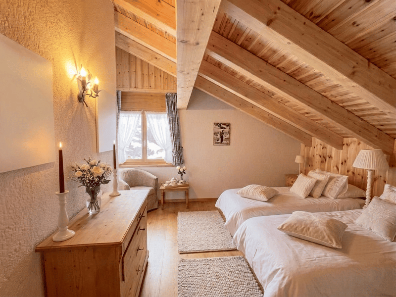 Bedroom with wooden beams, two beds, wooden dresser, two lamps, two candles, two vases with flowers, two rugs, two nightstands, picture frame, window with curtains, armchair, and a small table with a teapot and flowers.