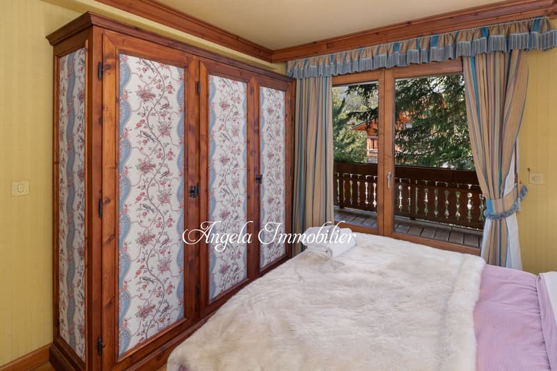 Spacious bedroom with wooden wardrobe, floral patterned curtains, and a balcony overlooking the trees outside