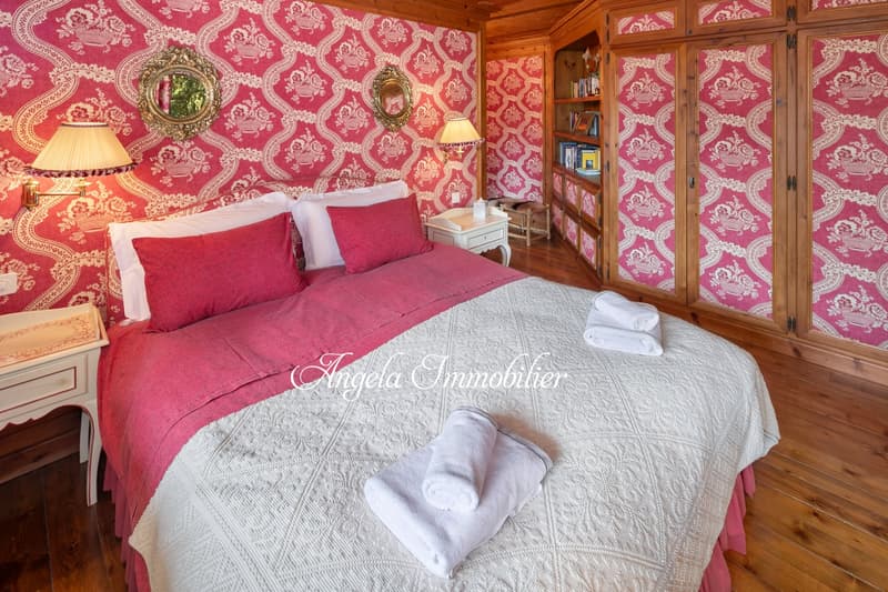 Spacious bedroom with ornate red and white patterned wallpaper, wooden furniture including a wardrobe, bedside tables, and a large bed with red bedding and a white quilt. The room also features decorative mirrors and a lamp.