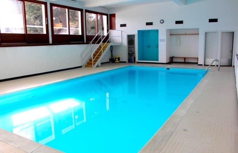 Indoor swimming pool, white walls, blue water, white tiles, large windows, stairs, benches, lockers