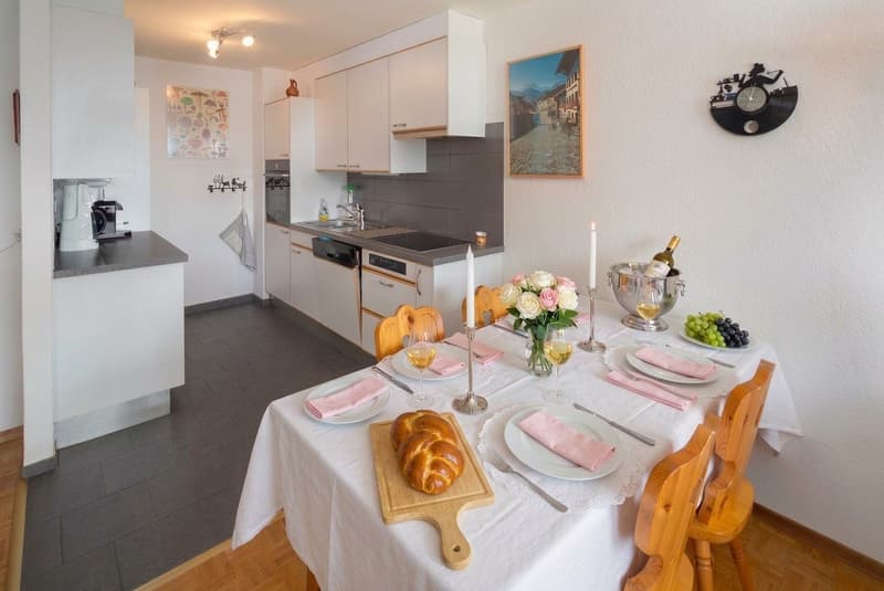Dining area with a table, chairs, wine glasses, flowers, bread, and kitchen appliances