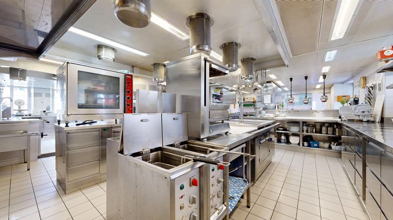 Commercial kitchen, tiled floor, stainless steel appliances, professional cooking equipment, double oven, extraction hoods, hanging utensils, various storage shelves, preparation tables, sinks, fryers.