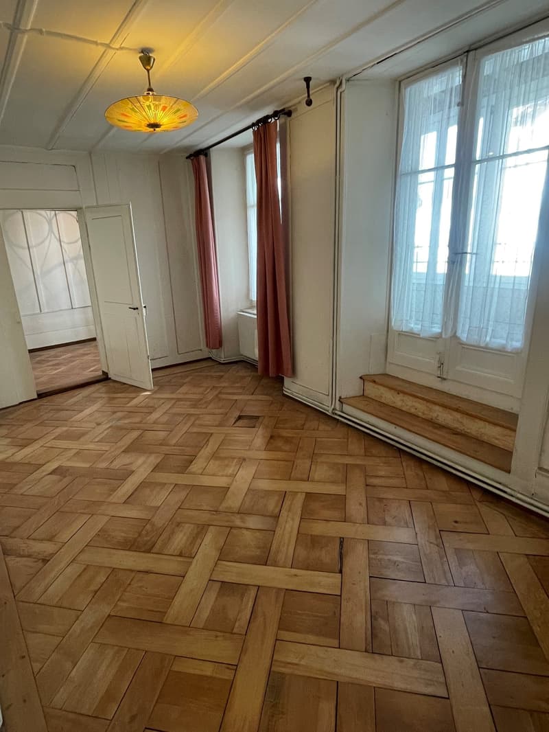 Wooden parquet flooring, white walls, two windows with curtains, an open door leading to another room, ceiling light fixture