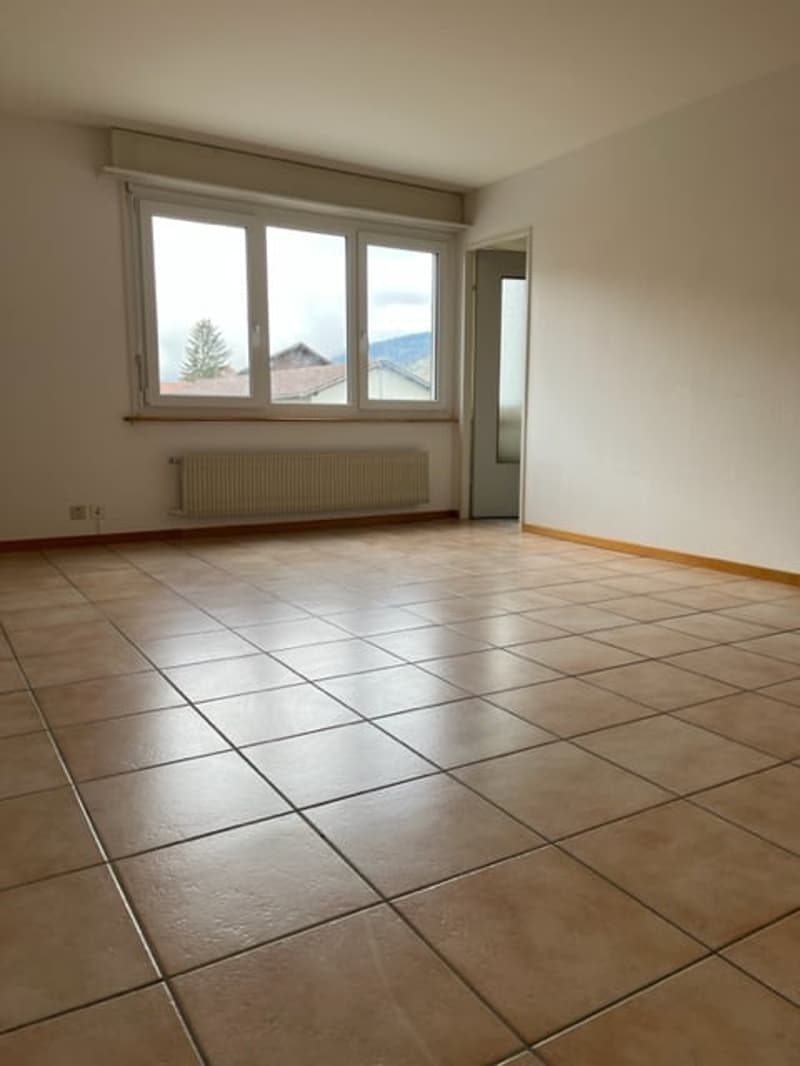An empty room with tiled flooring, white walls, a large window with a white radiator beneath it, and a door leading to another room.