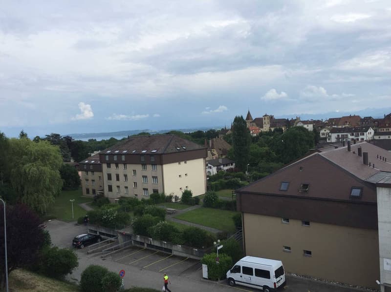 Aerial view of a residential building with a parking lot, surrounded by trees and greenery, with a distant view of mountains and a body of water.