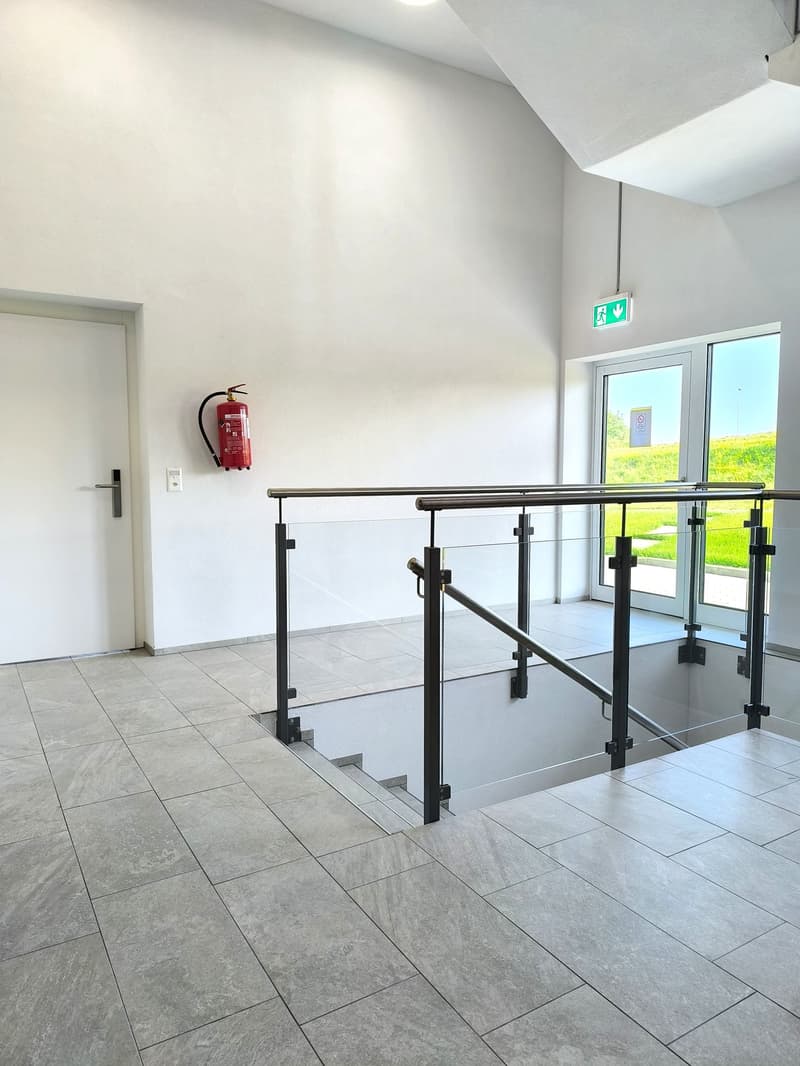 White walls, gray tile flooring, closed door, fire extinguisher, staircase, glass railing, emergency exit sign