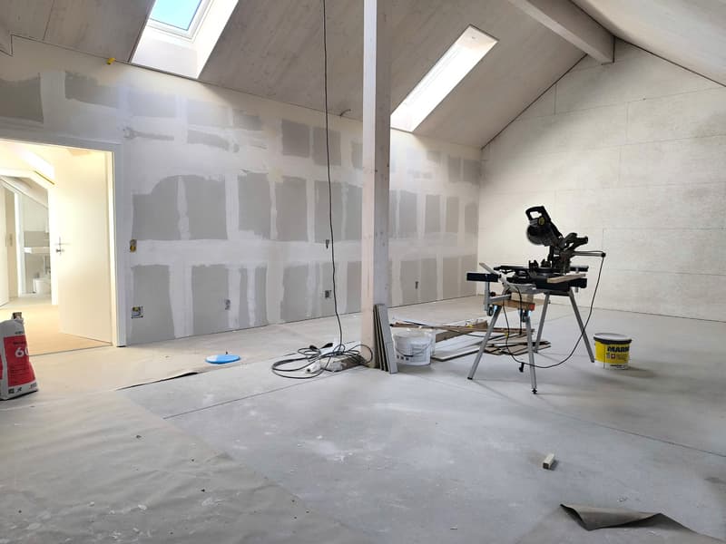 unfinished room, white walls, ceiling with skylights, wooden beams, electrical outlets, work bench, paint buckets, unfinished floor