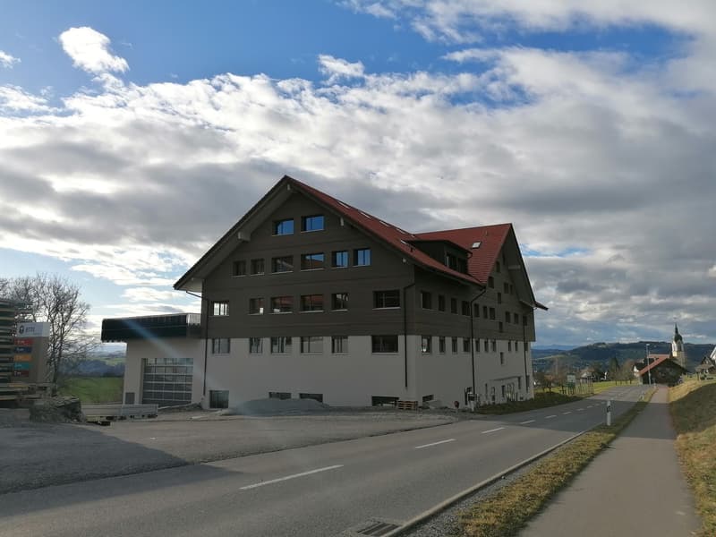 Multi-story building with brown and white exterior, red roof, garage, driveway, mountains and cloudy sky in the background.