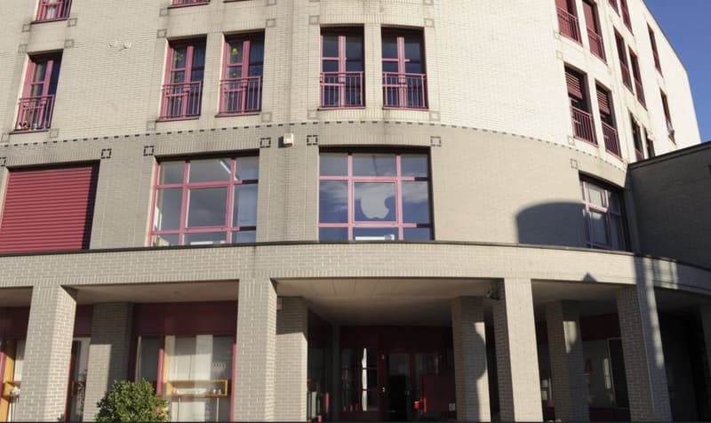 Modern building with brick facade, large windows, and red accents. The entrance has a double door with glass panels.