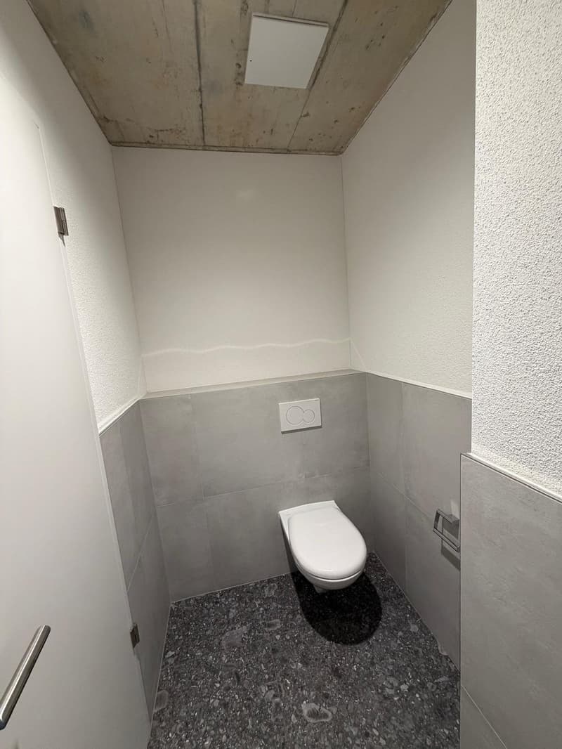 Small, modern, clean toilet room with white tile walls and a white floor, a white toilet, and a power outlet above it.