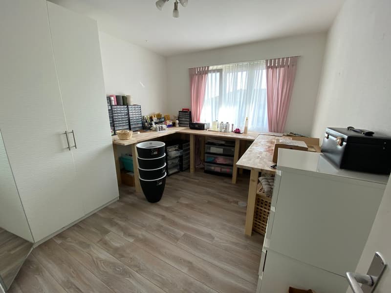 A room with a desk, cabinets, and a window with curtains. The room has a wooden floor and white walls. The desk has a black trash bin, a basket, and a storage box. The cabinets are white and have drawers. The window has pink curtains. There is a black bri