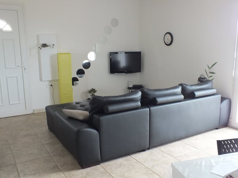 Spacious living room with a large black leather sofa, a mounted flat-screen TV, and decorative wall mirrors. There is also a yellow cabinet and a potted plant visible, suggesting a modern and minimalist decor style.