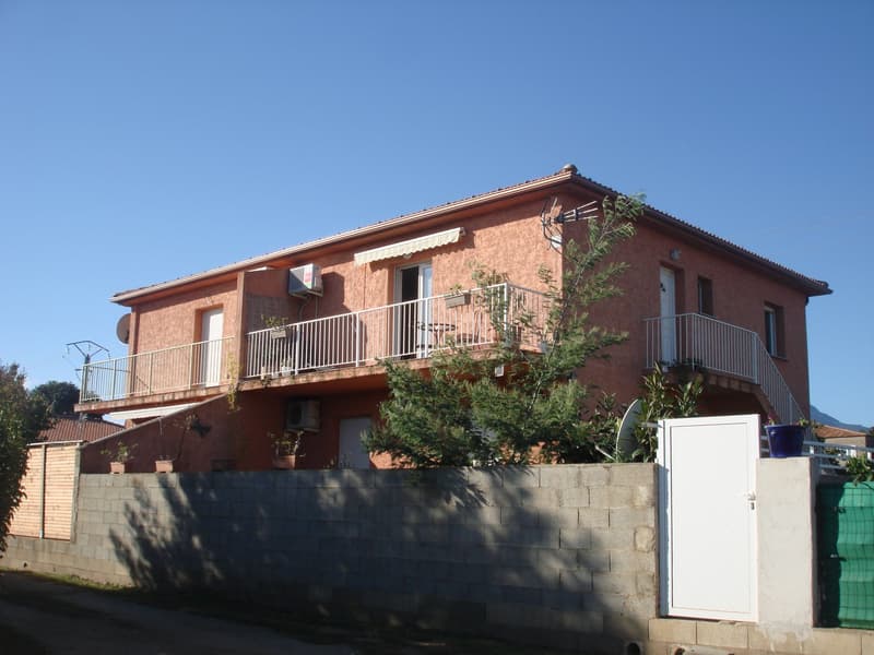 2-story house with a red-tiled roof, brown exterior walls, and balconies on the upper level. The house is surrounded by plants and trees.