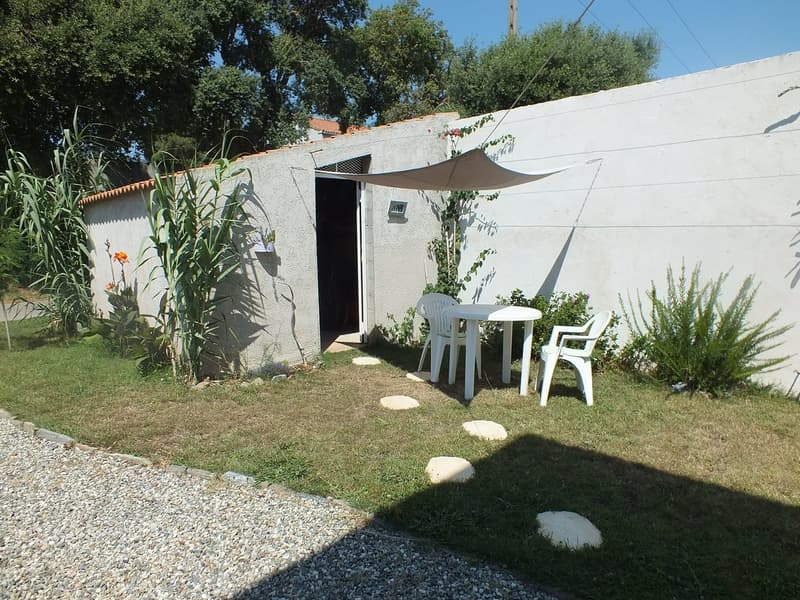 Small outdoor patio area with a white wall, a table and chairs, and a shade canopy. The space is surrounded by lush greenery, including plants and trees.