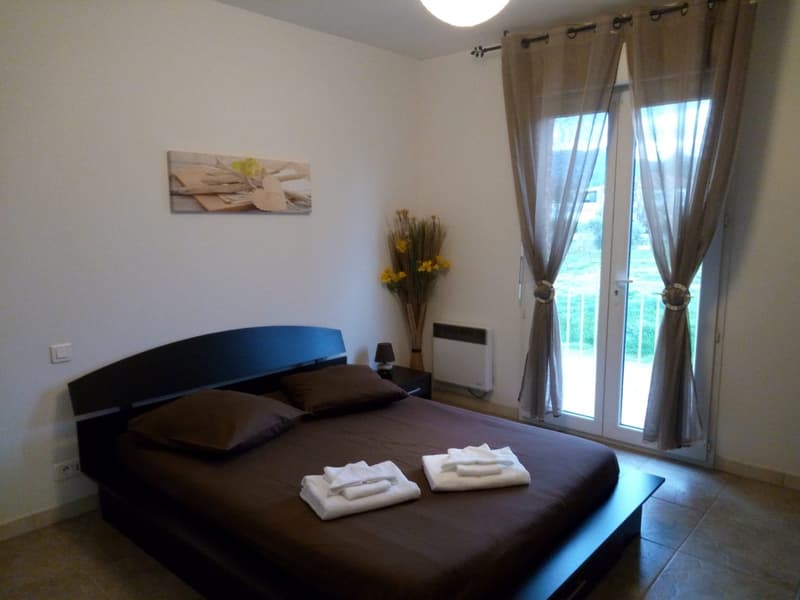 Spacious bedroom with a large bed, dark bedding, and a balcony or terrace visible through the window. The room has white walls, hardwood floors, and decorative elements like a floral arrangement and artwork on the wall.