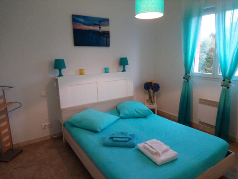 Bright and airy bedroom with white walls, a large window, a white dresser, and turquoise accents including bedding, lamps, and curtains. There is also a framed artwork on the wall and a vase of flowers on the dresser.