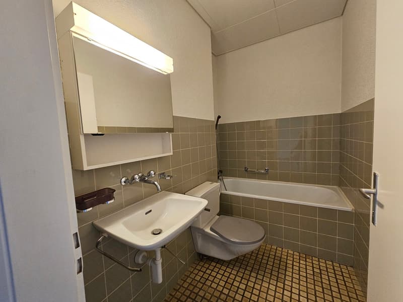 Bathroom with sink, toilet, bathtub, tiled wall and floor, a light over the mirror, and a silver soap dish.