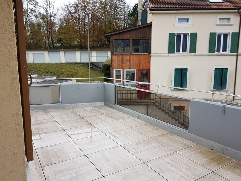 Balcony with tiled floor and glass railing, two buildings, one with brown roof and other with beige walls and green shutters, staircase, potted plant, and trees