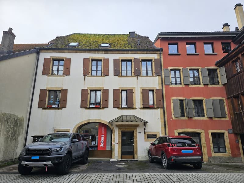 white building with brown shutters, green roof, multiple windows, two cars parked in front, two other buildings behind, brick walkways, sign in front