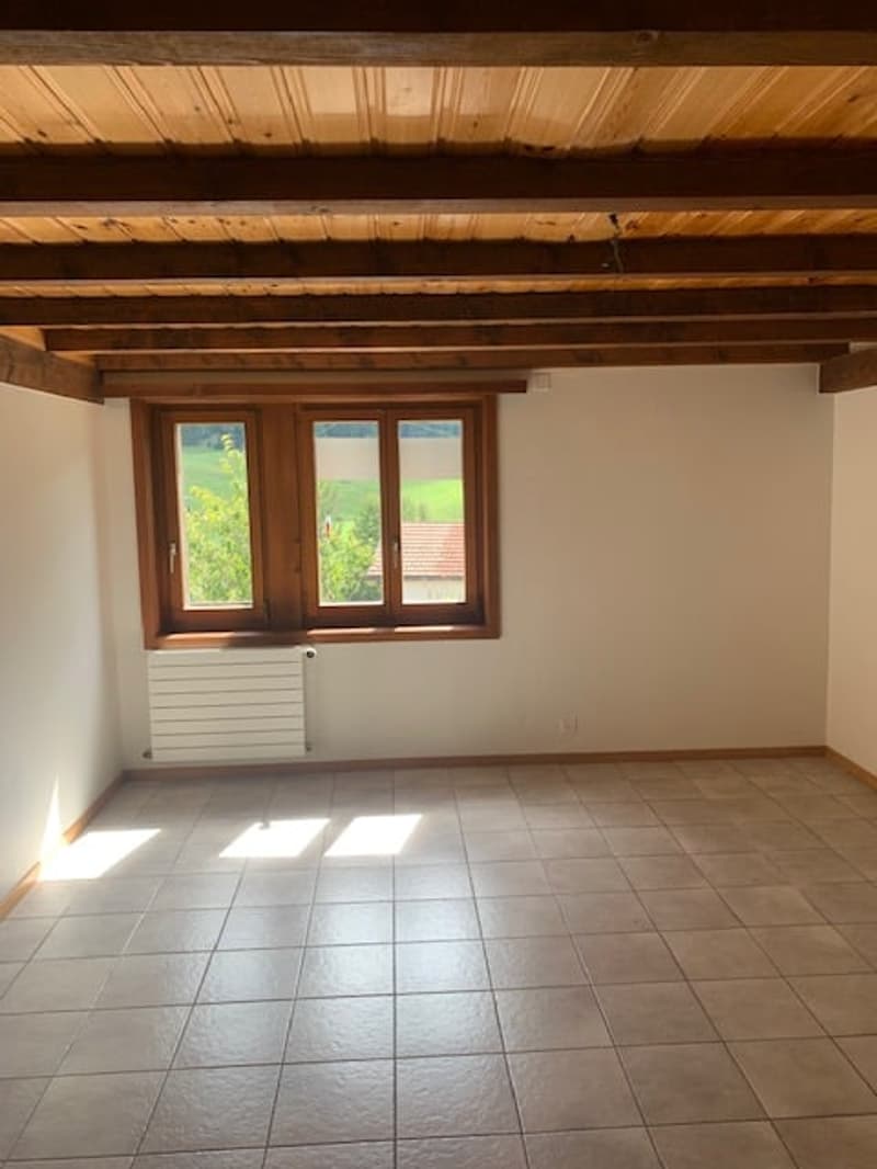 Empty room with wooden ceiling beams, white walls, and tile flooring. Large windows overlooking a scenic outdoor view.