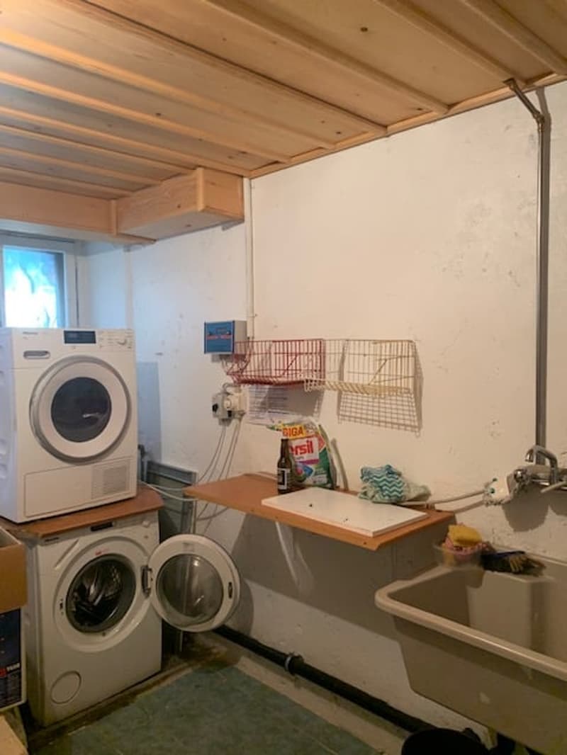 The image shows a laundry room with a washing machine and a dryer stacked on top of each other. There is also a sink and a counter space with some storage items on it. The room has a wooden ceiling and walls.