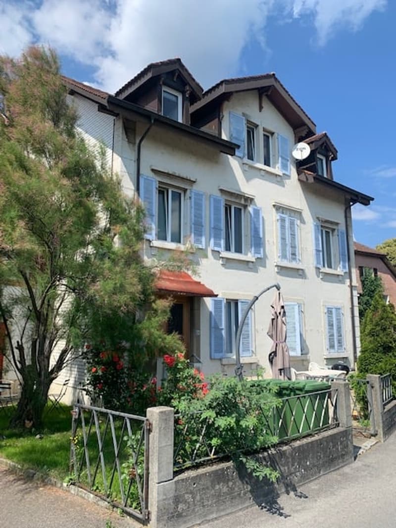 Two-story house with white exterior, blue shutters, and a balcony on the second floor. The house has a garden in the front with a fence and flowers.