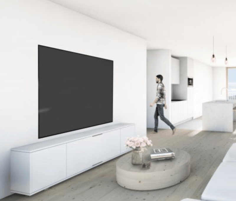 Large flat-screen TV mounted on the wall, white TV stand, round concrete coffee table, hardwood flooring, open floor plan with visible hallway in the background
