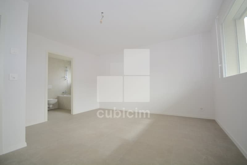 Spacious room with white walls, large windows, and a bathroom visible through the doorway. The room has a minimalist, clean design with a concrete floor.