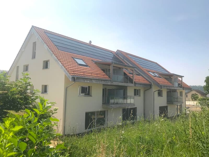 3-story apartment building with red tile roof, white exterior walls, and several balconies. The building has solar panels installed on the roof. There appears to be a parking area in front of the building surrounded by greenery and vegetation.