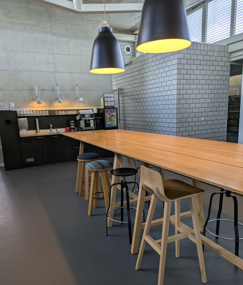 Modern kitchen with concrete walls, white subway tile backsplash, black cabinets, wooden countertop, and large pendant lights with yellow interiors. The kitchen also features a dishwasher and a long wooden dining table with stools.