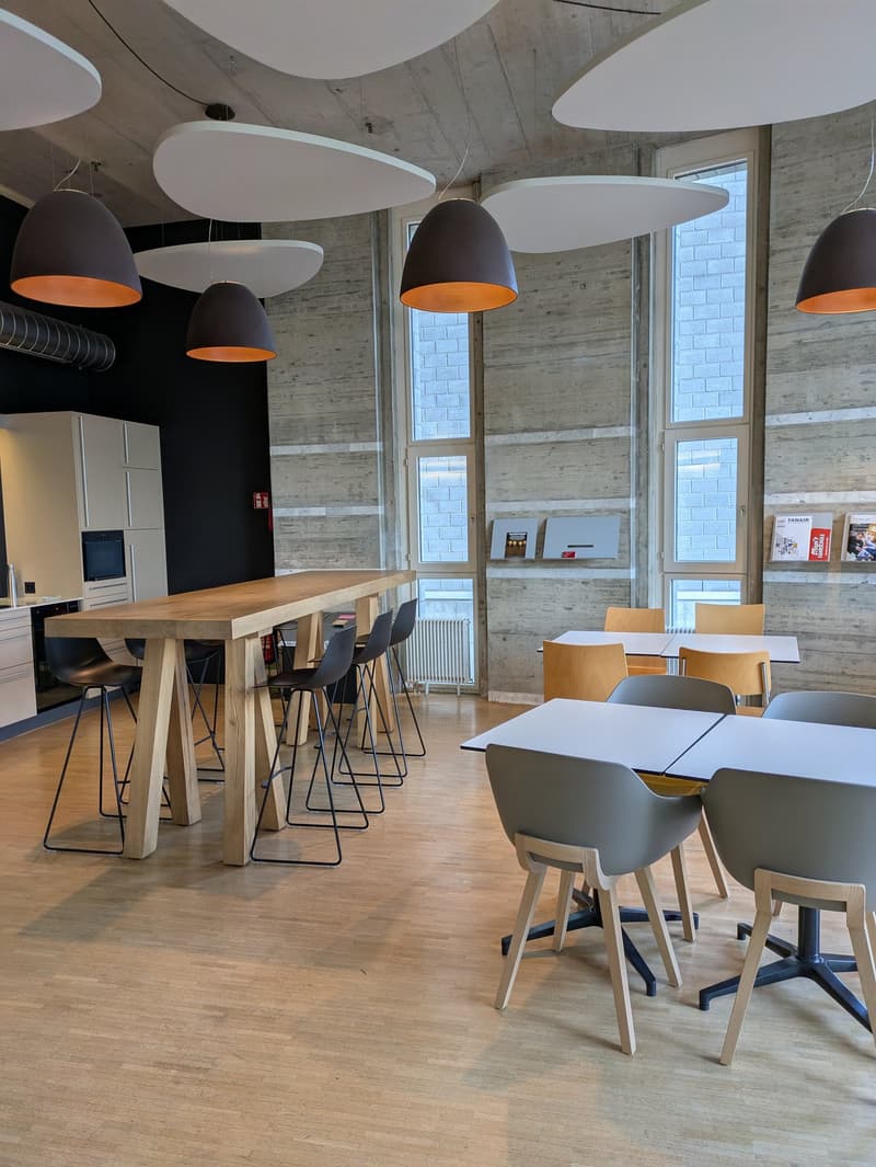 The image shows a modern and open office or workspace with concrete walls, large windows, and a mix of wooden and gray furniture. There are several round pendant lights hanging from the ceiling, and the space includes a long wooden table with stools, as w