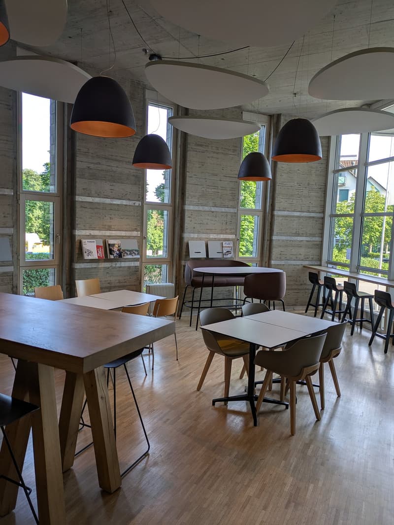 The image shows a modern and spacious cafe or restaurant interior. The space features large windows that provide natural light and views of the surrounding greenery outside. The walls are made of exposed concrete, giving the space an industrial and minima