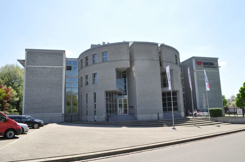 A large, multi-story gray brick building with a curved facade and large windows. There is a paved area in front of the building with some cars parked. The building appears to be a commercial or institutional structure.