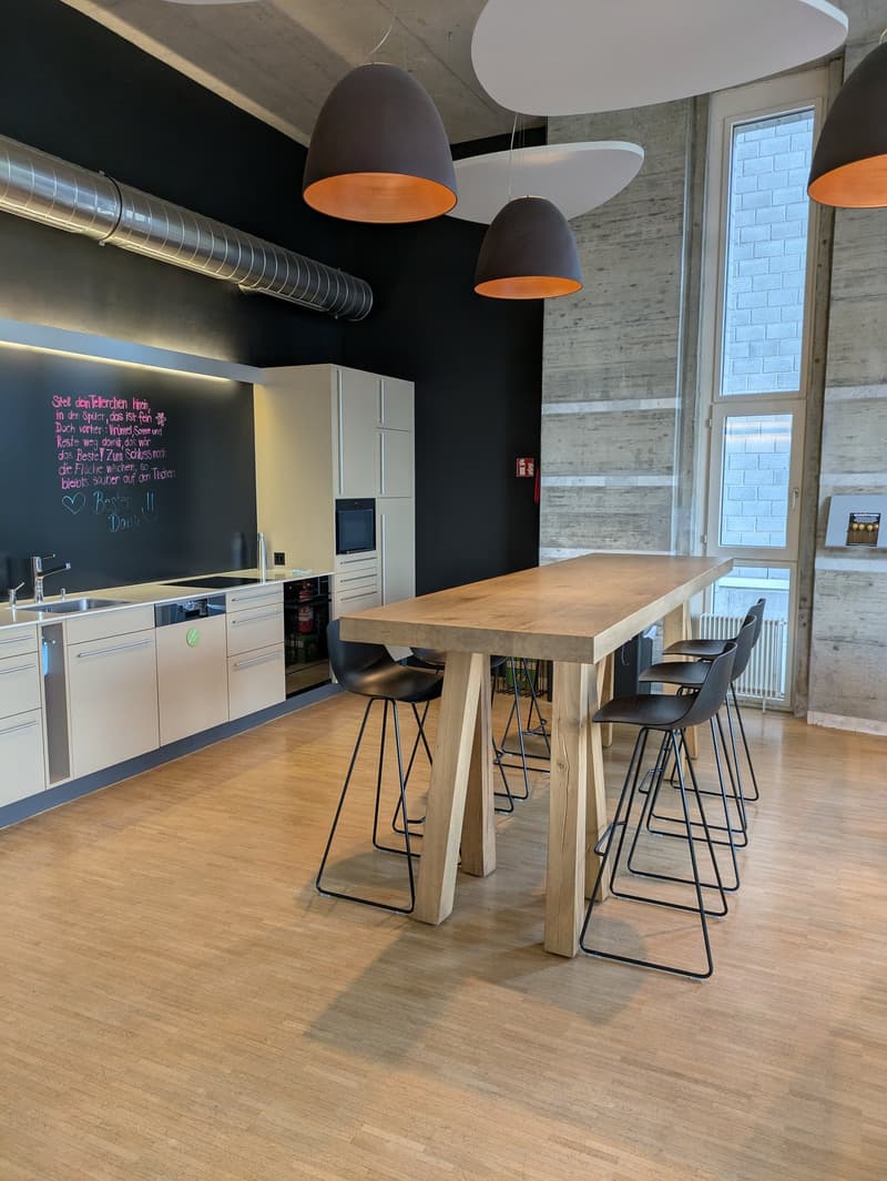 Modern kitchen with wooden dining table and black bar stools, black chalkboard wall, pendant lighting, and stainless steel appliances including a dishwasher.
