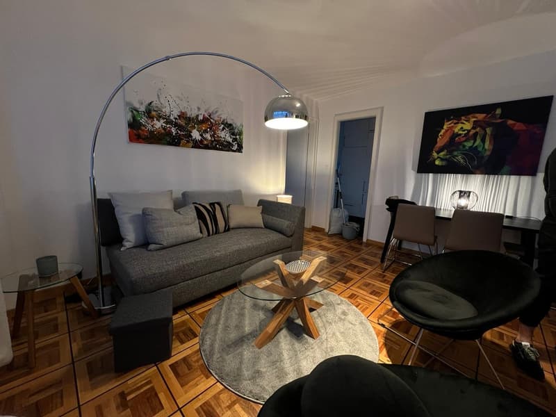 Living room with gray couch, glass coffee table, round rug, floor lamp, two armchairs, desk, and paintings on the wall.