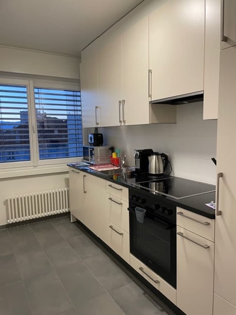 Kitchen. Modern kitchen with white cabinets, black countertops, and stainless steel appliances. Features an oven, coffee maker, and a view of the city through large windows.