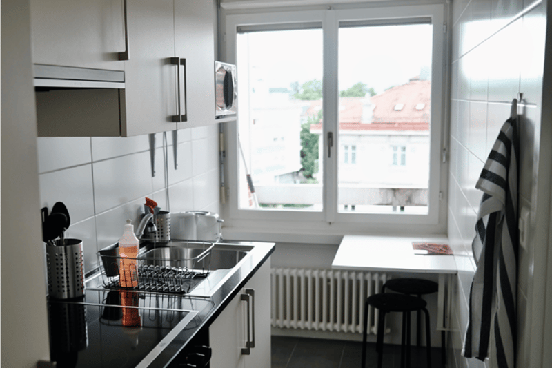 Kitchen. Modern kitchen with white cabinets, stainless steel appliances, a sink with a dish rack, a microwave, a stove, and a small table with a stool. The room has large windows offering a view of the city.