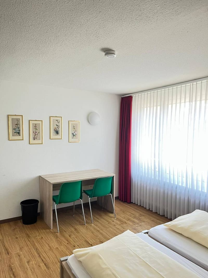 Bedroom with hardwood flooring, white walls, framed artwork, a desk with green chairs, and a large window with red curtains.
