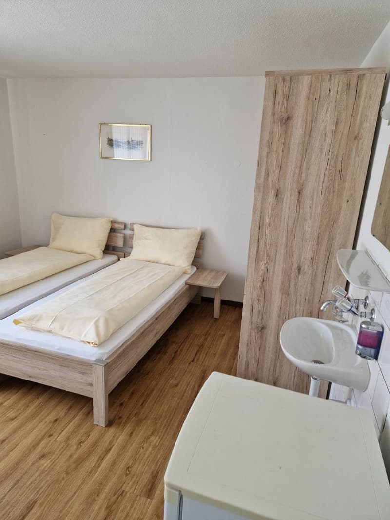 Spacious bedroom with wooden furniture, including a bed with beige bedding, a side table, and a wardrobe. The room also features a small table and a sink, suggesting it may be a studio or efficiency apartment. The overall design has a minimalist and moder