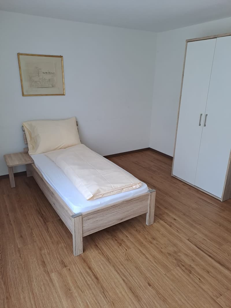 Bedroom with a single bed, wooden nightstand, and a white wardrobe. The room has hardwood flooring and white walls with a framed artwork on the wall.