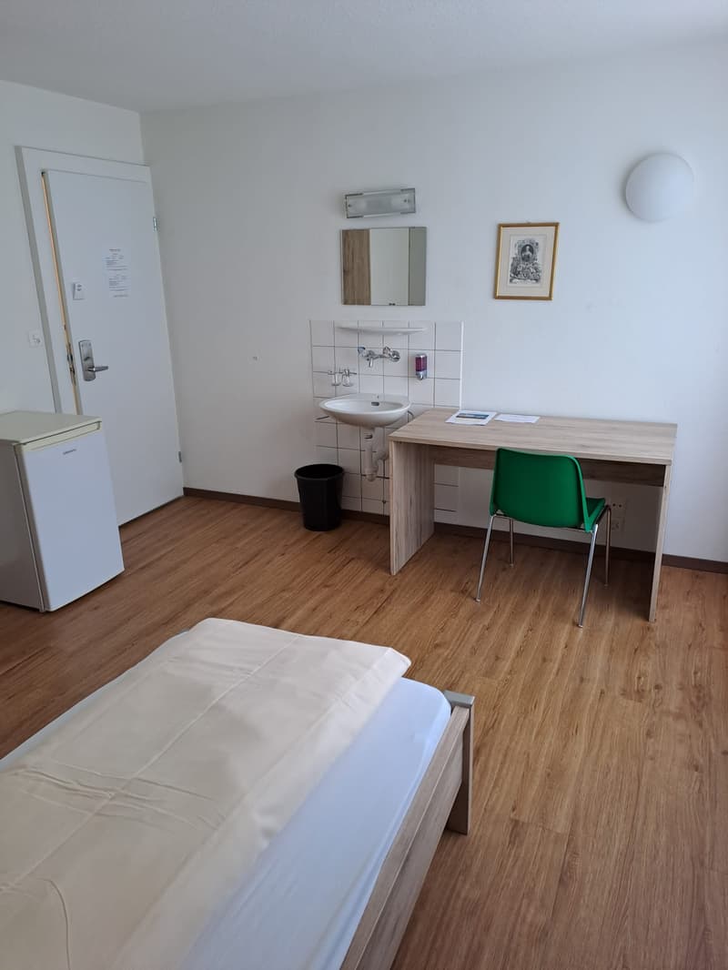 The image shows a studio apartment with a bed, a desk with a chair, a bathroom vanity with a sink, and a small refrigerator. The room has white walls, a hardwood floor, and some decor elements like a framed artwork on the wall.