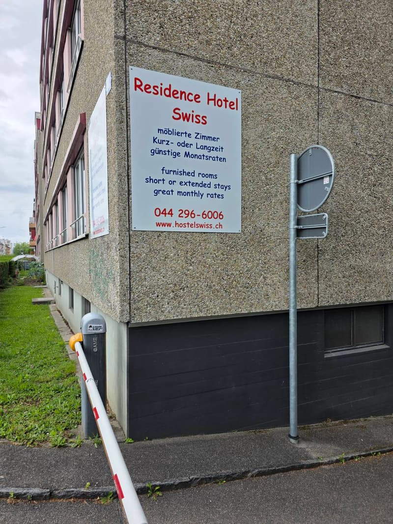 Exterior view of a building with a sign reading "Residence Hotel Swiss" advertising furnished rooms for short or extended stays with great monthly rates. The building has a textured stone facade and a black garage door.